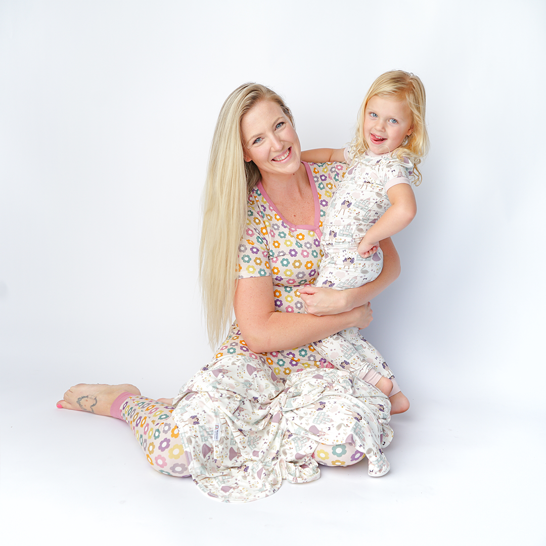 a mom sits on the floor with her daughter. the daughter smiles with her hand on her hip. she wears the "once upon a time" 2-piece matching pajama set. the "once upon a time" print is a mix of pinks, purples, whites, blues, and yellows. you can see fairy princess and regular princess, unicorns, swans, and castles. theres also hearts, stars and sparkles scattered around the print. 