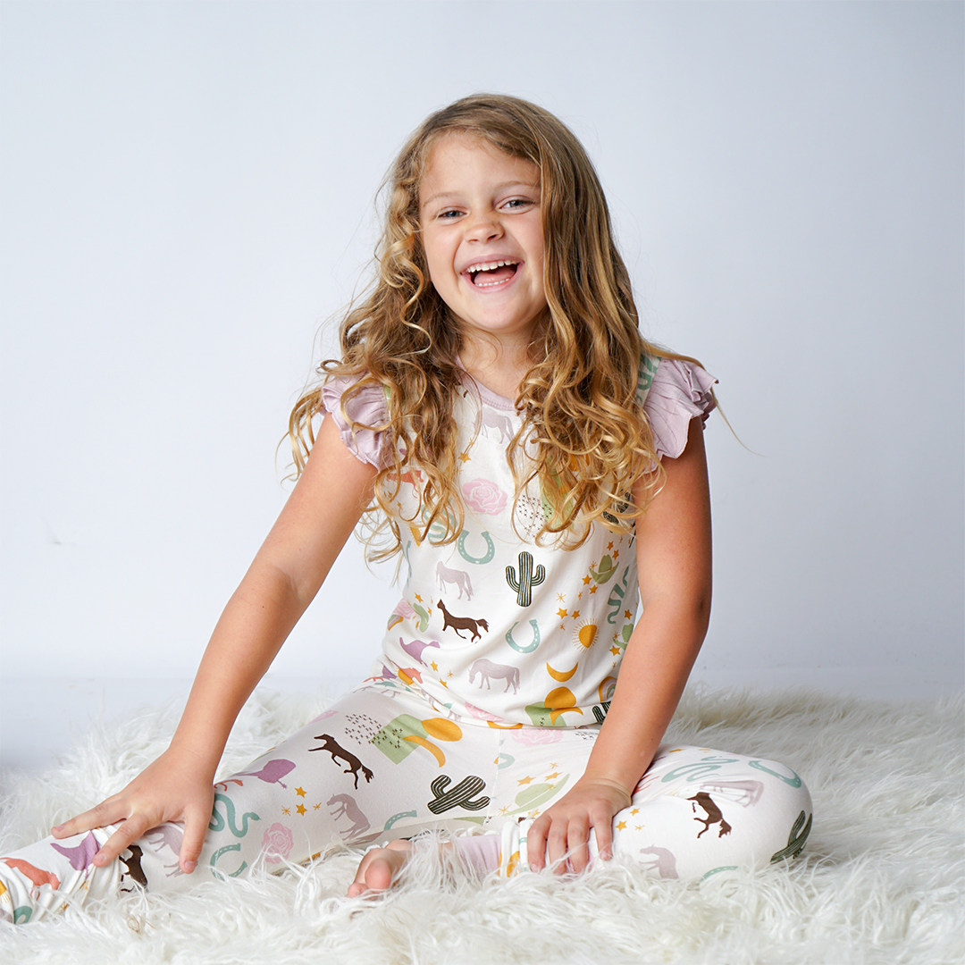 young girl laughing. she wears the "wild and free" matching pajama set. the "wild and free" print is a mix of colorful horses, snakes, cowboy hats, cacti, suns, stars, moons, and rainbows, all on a white background. this is a boho cowboy design. 