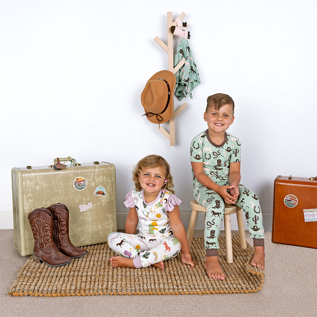 brother and sister smile at the camera. the girl wears the "wild and free" matching pajama set. the "wild and free" print is a mix of colorful horses, snakes, cowboy hats, cacti, suns, stars, moons, and rainbows, all on a white background. this is a boho cowboy design. 