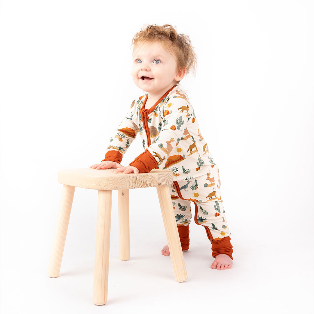 a baby stands holding on to a wooden stool for balance. the baby is wearing the "desert friends" convertible. the "desert friends" print is a scattered pattern of armadillo, cacti, other desert plants, rocks, desert foxes, lizards, and desert birds. this is all on a beige background. 