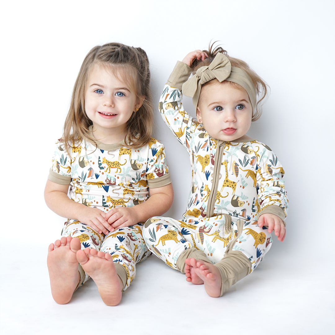 2 girls sit on the floor. the older girl on the left wears the "jungle friends" matching set. the younger girl wears the "jungle friends" convertible. the "jungle friends" print is a mix of jungle animals that include giraffes, lions, cheetahs, sloths, snakes, and forest plants. 