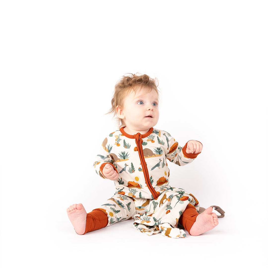 baby sits on the floor looking to something to the right of him. he is wearing the "desert friends" convertible and the "desert friends" lovey is draped on his leg. the "desert friends" print is a scattered pattern of armadillo, cacti, other desert plants, rocks, desert foxes, lizards, and desert birds. this is all on a beige background. 