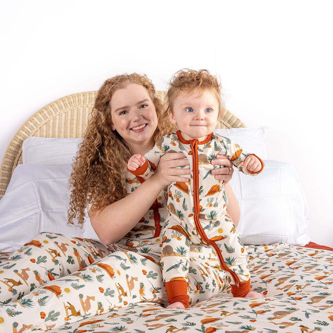 and mom and baby pose themselves on a bed. the baby is being held up by the mom. the baby wears the "desert friends" convertible. the mom is in the "desert friends" women's top. the "desert friends" print is a scattered pattern of armadillo, cacti, other desert plants, rocks, desert foxes, lizards, and desert birds. this is all on a beige background. 