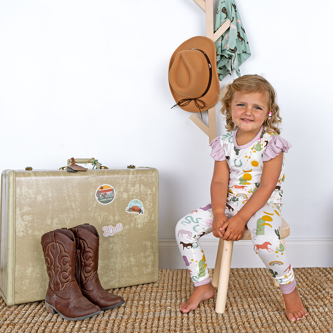 a young girl sits on a wooden stool and smiles. she is wearing the "wild and free" matching pajama set. there is a western themed set up around her, that includes an old fashion suitcase, cowboy boots, and a cowboy hat. the "wild and free" print is a mix of colorful horses, snakes, cowboy hats, cacti, suns, stars, moons, and rainbows, all on a white background. this is a boho cowboy design. 