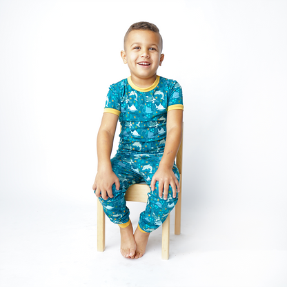 boy sits in a wooden chair wearing his "ocean friends" matching 2-piece set. the "ocean friends" print is a combination of dolphins, stingrays, fish, starfish, coral, bubbles, and sharks, all spread out on a deep sea blue background.