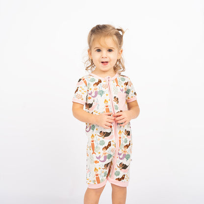 girl poses in her "making waves" shortie. the "making waves" print has a diverse spread of mermaids, sea coral, starfish, fish, and bubbles all spread out in different colors. this is all put on a beige background.