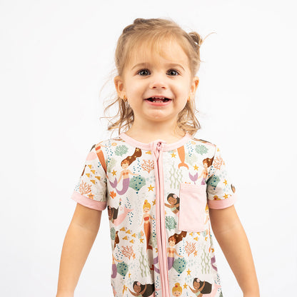 girl smiles wearing her "making waves" shortie. the "making waves" print has a diverse spread of mermaids, sea coral, starfish, fish, and bubbles all spread out in different colors. this is all put on a beige background.