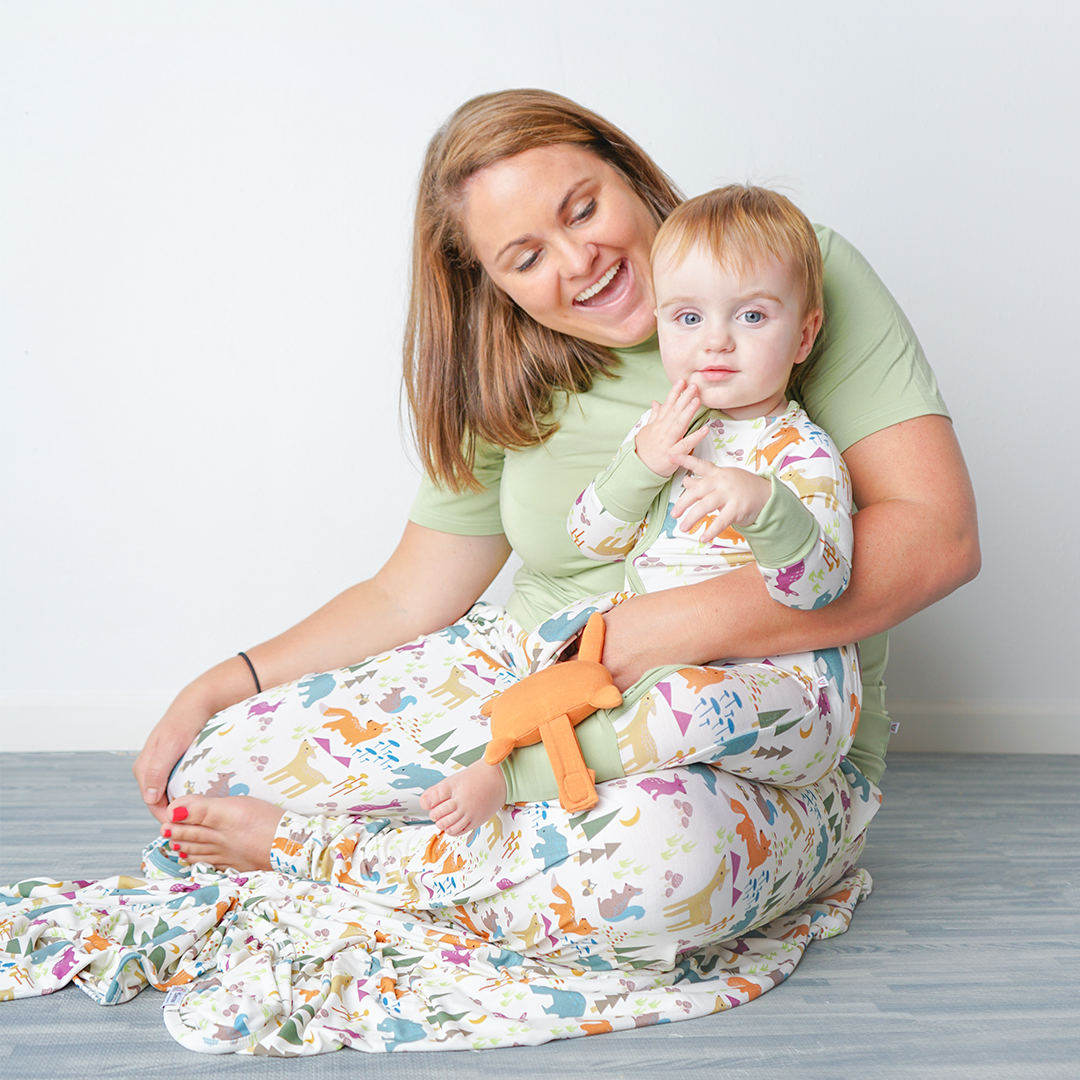 mom and baby sit together wearing their "forest friends" apparel.  mom wears the relaxed pants and the baby wears the convertible and hold the lovey. the "forest friends" print has a mix of red foxes, brown deer, blue bears, purple bunnies, trees, grass, mushrooms, acorns, moons, and other colored dots scattered over a white background. 