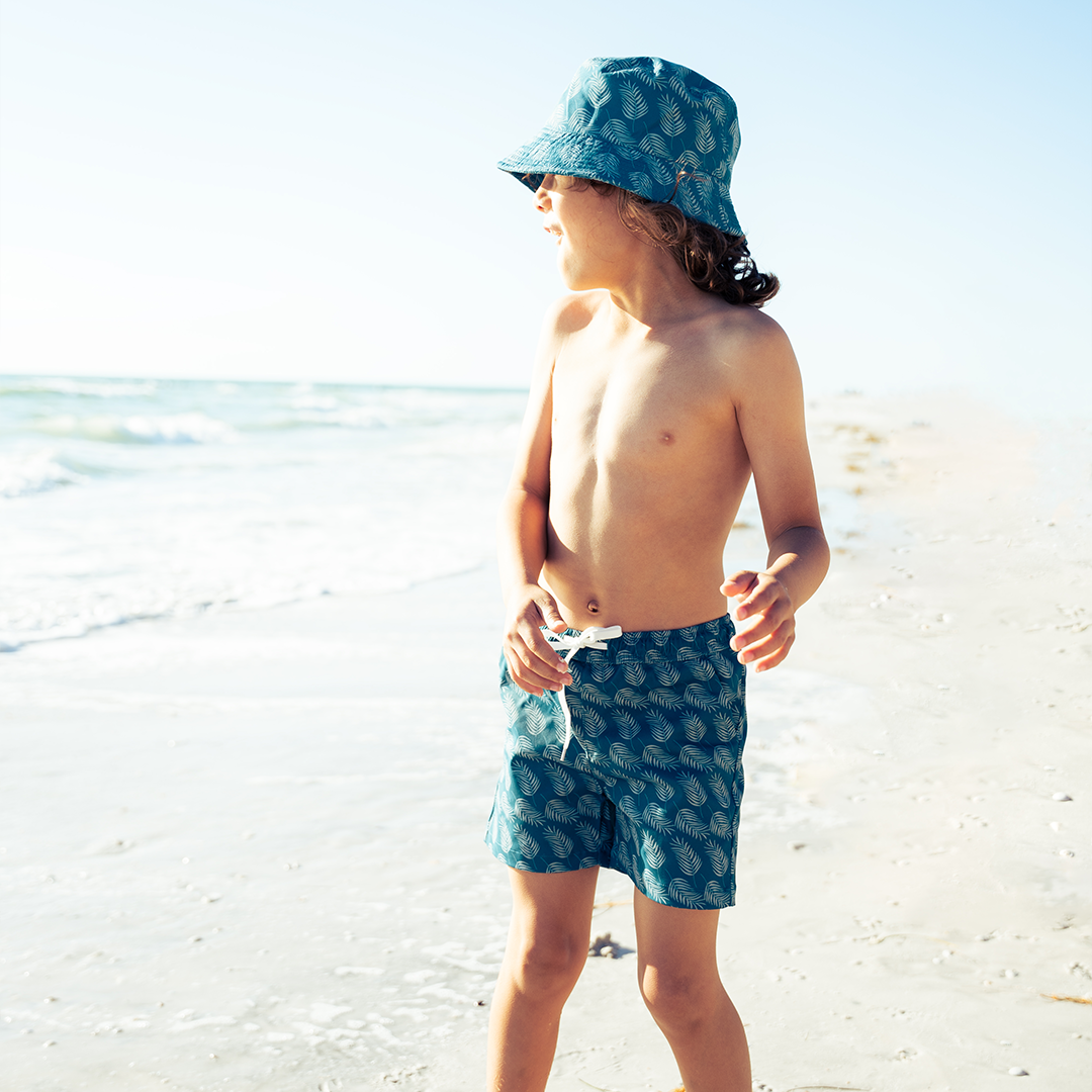 a boy looks at the ocean as he stands on the beach. he wears the "palms in paradise" swim trunks and bucket hat. the "palms in paradise" print is a pretty dark ocean blue background with palms in a lighter blue spread out along the print.