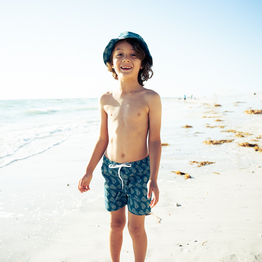 a boy smiles and stands on the beach. he is wearing the "palms in paradise" swim trunks. the "palms in paradise" print is a pretty dark ocean blue background with palms in a lighter blue spread out along the print. the "palms in paradise" print is a pretty dark ocean blue background with palms in a lighter blue spread out along the print.