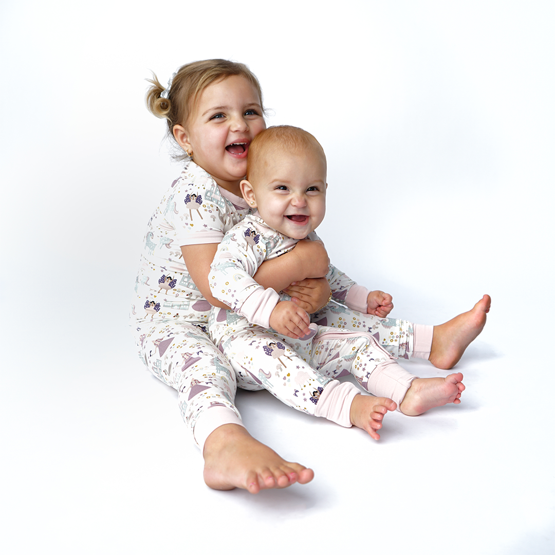 2 sisters sit on the floor. the older sister is in the "once upon a time" matching pajama set. she holds her baby sister in her lap. the baby wears the "once upon a time" convertible. the "once upon a time" print is a mix of pinks, purples, whites, blues, and yellows. you can see fairy princess and regular princess, unicorns, swans, and castles. theres also hearts, stars and sparkles scattered around the print. 