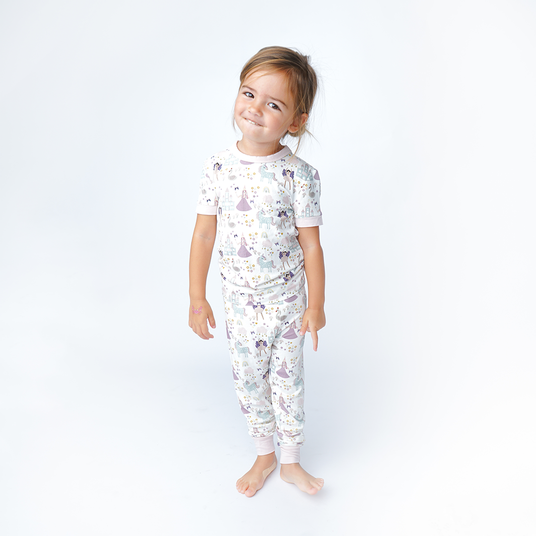 a girl poses and smiles in her "once upon a time" matching pajama set. the "once upon a time" print is a mix of pinks, purples, whites, blues, and yellows. you can see fairy princess and regular princess, unicorns, swans, and castles. theres also hearts, stars and sparkles scattered around the print. 