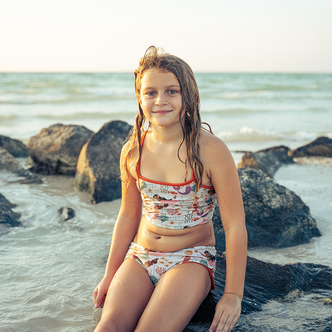 young girl sits on a rock in the ocean. she is smiling at the camera and wears the "making waves" reversible bikini. there are more rocks behind her and ocean waves behind her. the "making waves" print has a diverse spread of mermaids, sea coral, starfish, fish, and bubbles all spread out in different colors. this is all put on a beige background.