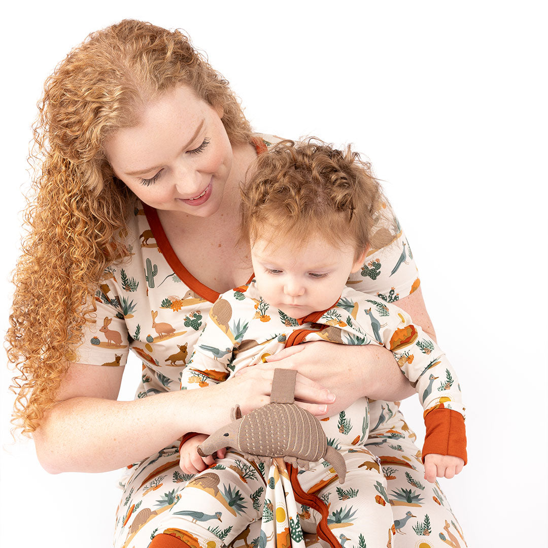 a baby sits on a mothers lap. they are in matching "desert friends" prints. the mom is wearing the women's top and the baby is in the convertible. they are both holding and looking at the "desert friends" lovey. the "desert friends" print is a scattered pattern of armadillo, cacti, other desert plants, rocks, desert foxes, lizards, and desert birds. this is all on a beige background. 