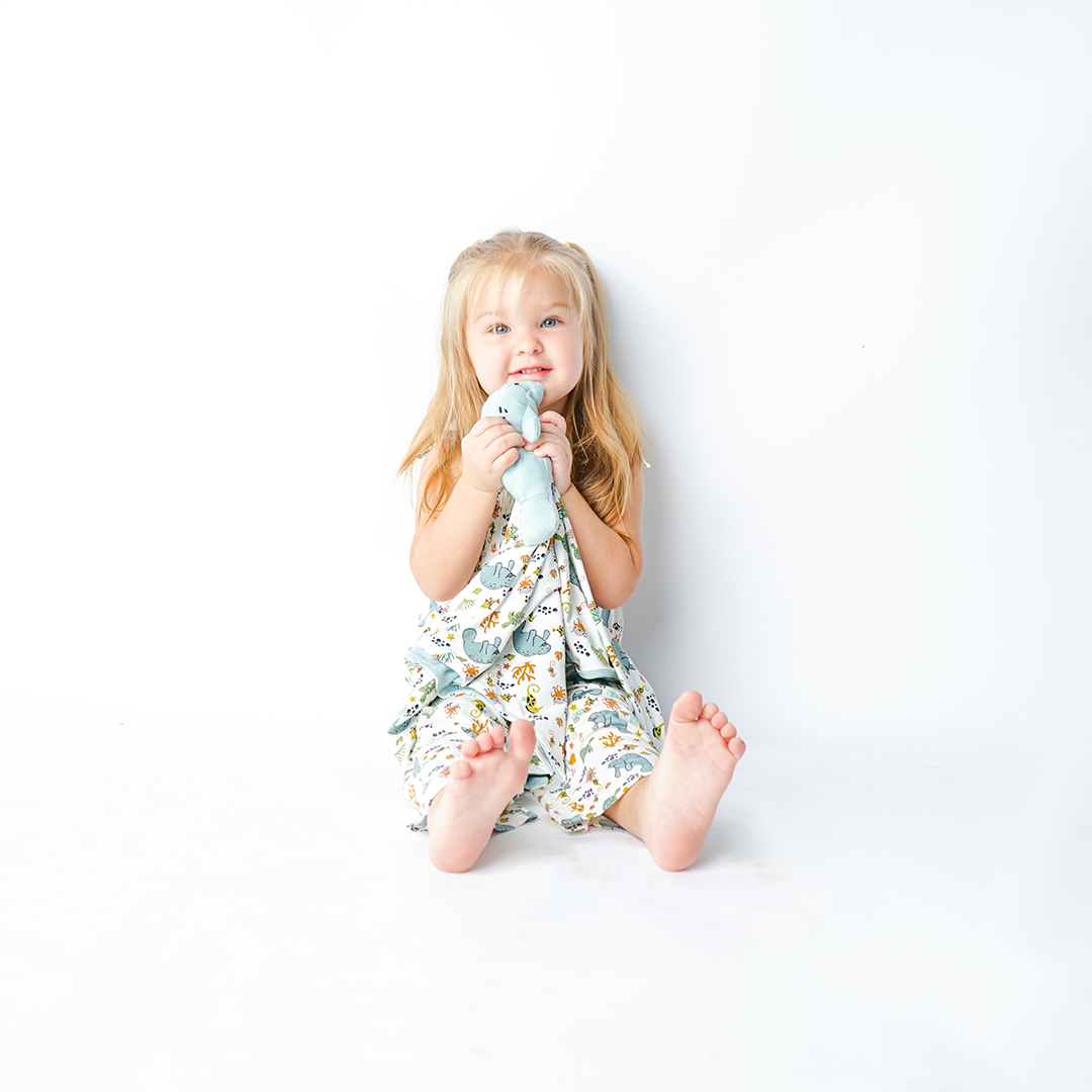 small toddler girl covers herself with the "manatee" lovey. the print "manatee" unites the florida sea life in a majestic print. there is a variation of florida manatees, yellow and pink sea horses, yellow and pink starfish, bubbles, turtles, and coral patterned across a white background