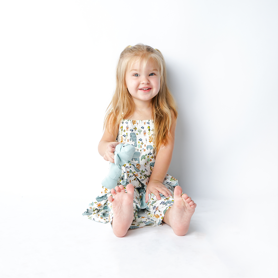 small toddler girl sits with the "manatee" sundress and the "manatee" lovey. the print "manatee" unites the florida sea life in a majestic print. there is a variation of florida manatees, yellow and pink sea horses, yellow and pink starfish, bubbles, turtles, and coral patterned across a white background