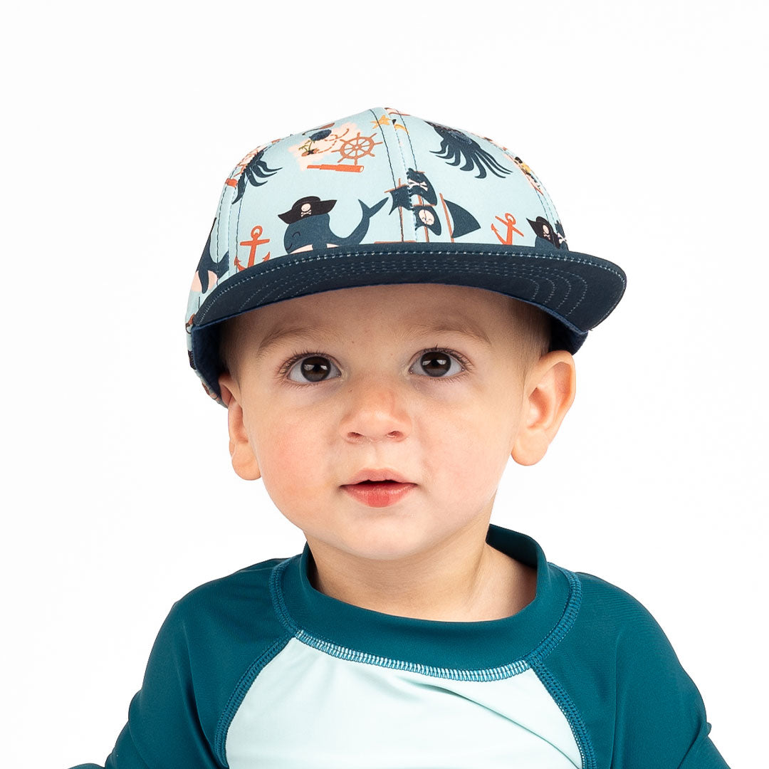 baby looks into the camera. he is wearing the "pirate's life" snapback hat. the "pirate's life" print is a mix of octopus, whales, and seagulls all wearing pirate hats. there are also anchors, pirate ships, treasure maps, and starfish mixed into the pattern. this is all displayed on a teal background. 