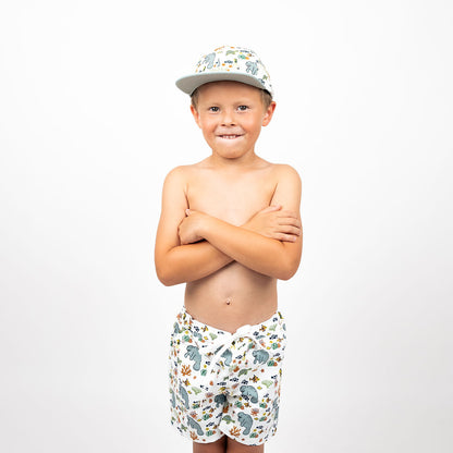 a boy stands with his arms crossed. he is wearing the "manatee" boy swim trunks and the matching snapback hat. the print "manatee" unites the florida sea life in a majestic print. there is a variation of florida manatees, yellow and pink sea horses, yellow and pink starfish, bubbles, turtles, and coral patterned across a white background