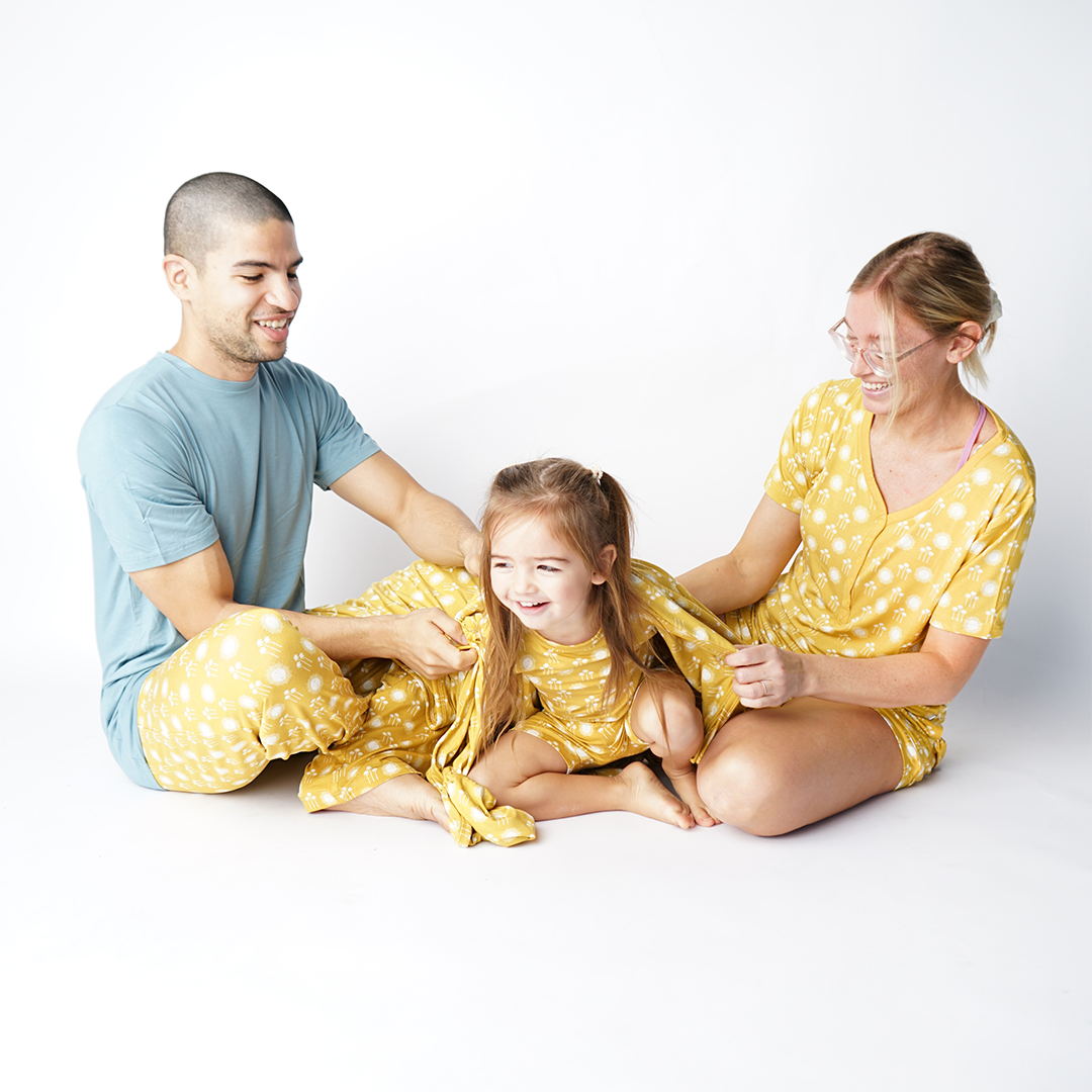 a family of 3 smiling. the mom and the baby are both wearing the sunny days print. the father is wearing the sunny days  bottoms and the "blue surf" unisex tee shirt. 