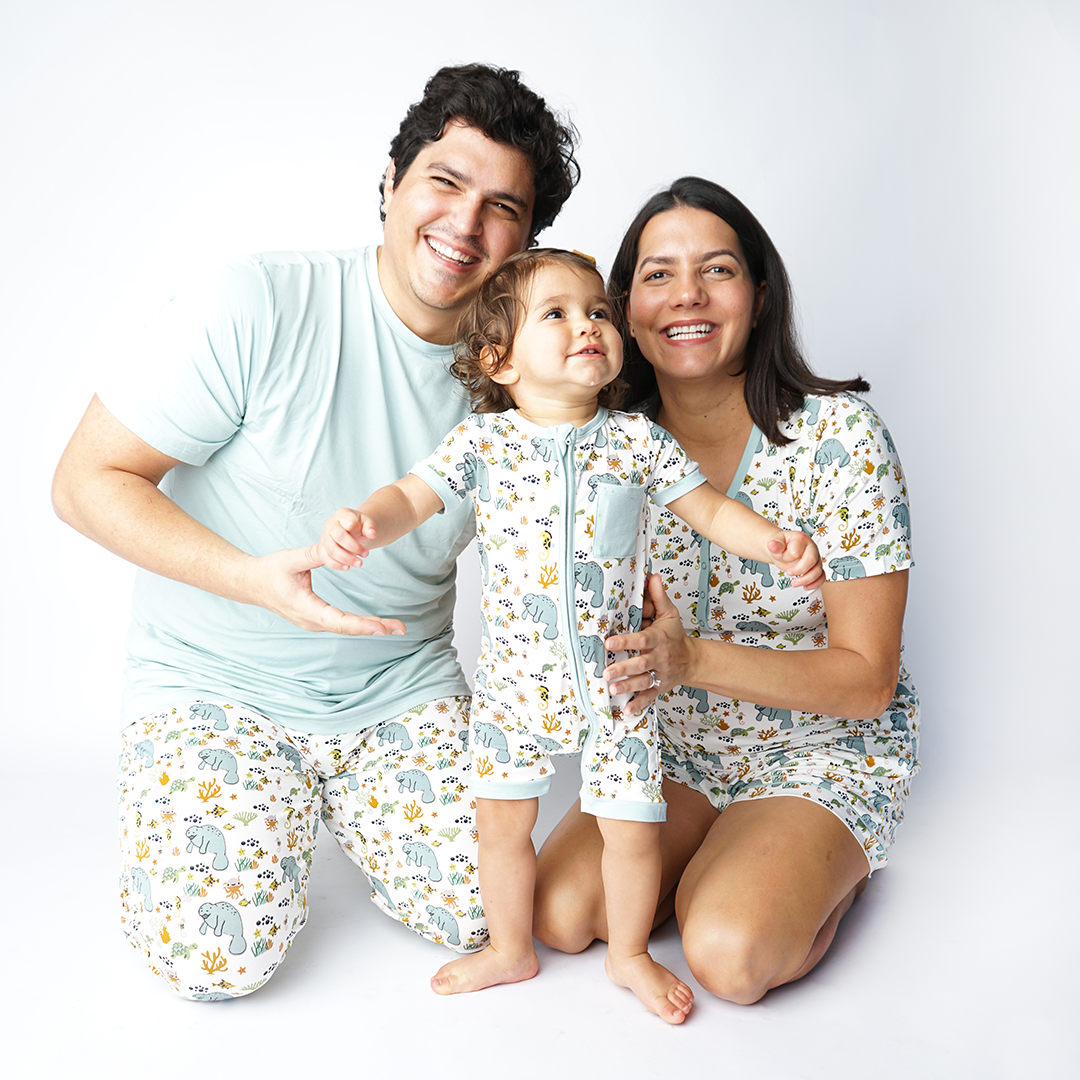 a family of 3 smiling. the mom and the baby are both wearing the manatee print. the father is wearing the manatee bottoms and the "blue surf" unisex tee shirt. 