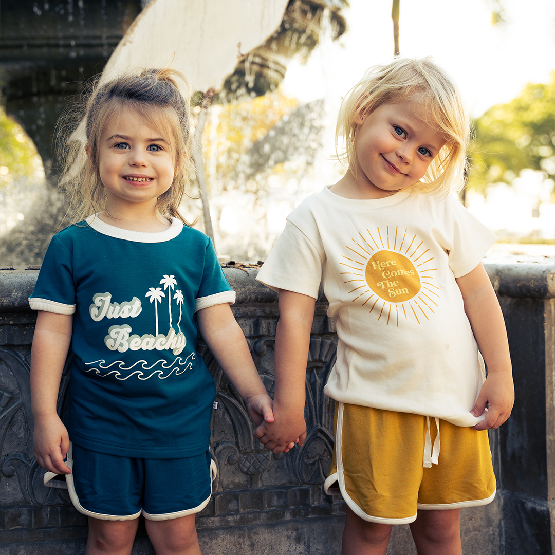 2 girls stand up against a water fountain. they hold hands. the girl on the left is wearing the blue terry shorts and the "just beachy" tee. the girl on the left wears the "golden sunshine bamboo terry track shorts" and the "here comes the sun" tee. 