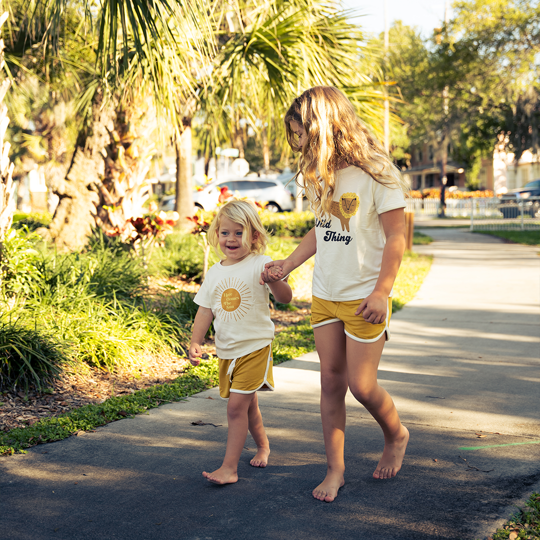 2 girls walk on a sidewalk hand in hand. they are wearing the golden sunshine bamboo terry track shorts. the younger one is wearing the "here comes the sun" tee. the older one is wearing the "wild things" tee. they are outside and you can see grass, palm trees, and a parking lot behind them. 