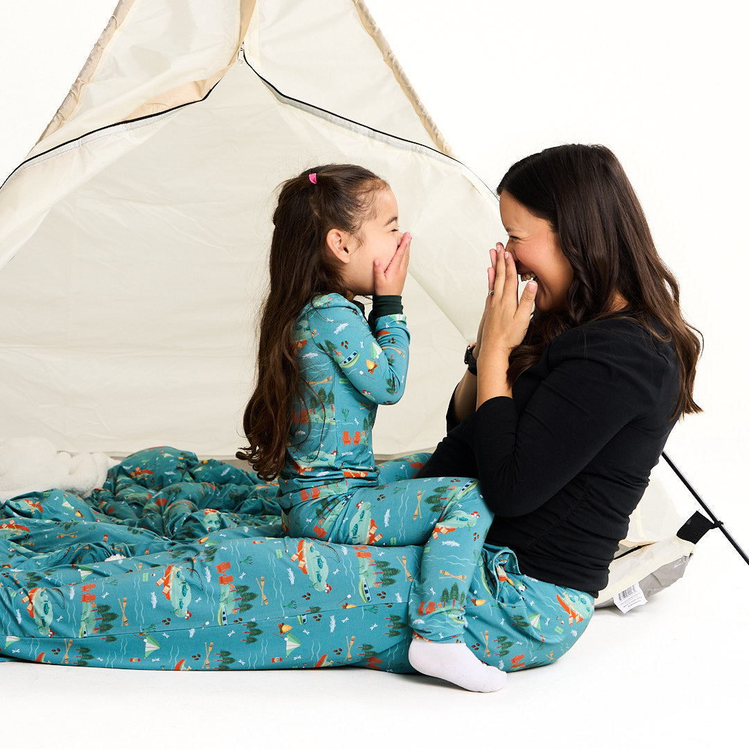 mom and daughter sit under a tent. they look at one another, laughing. they cover their faces with their hands. the daughter wears the "jump in the lake" matching pajama set. the mom wears the "jump in the lake" relaxed pants. the "jump in the lake" pattern is a print to capture the day in the life on a lake. you can fine fishing poles, tents, camp fires, adirondack chairs, marsh mellows, ores, canoes, forest trees, and different types of fish and fishing gear.