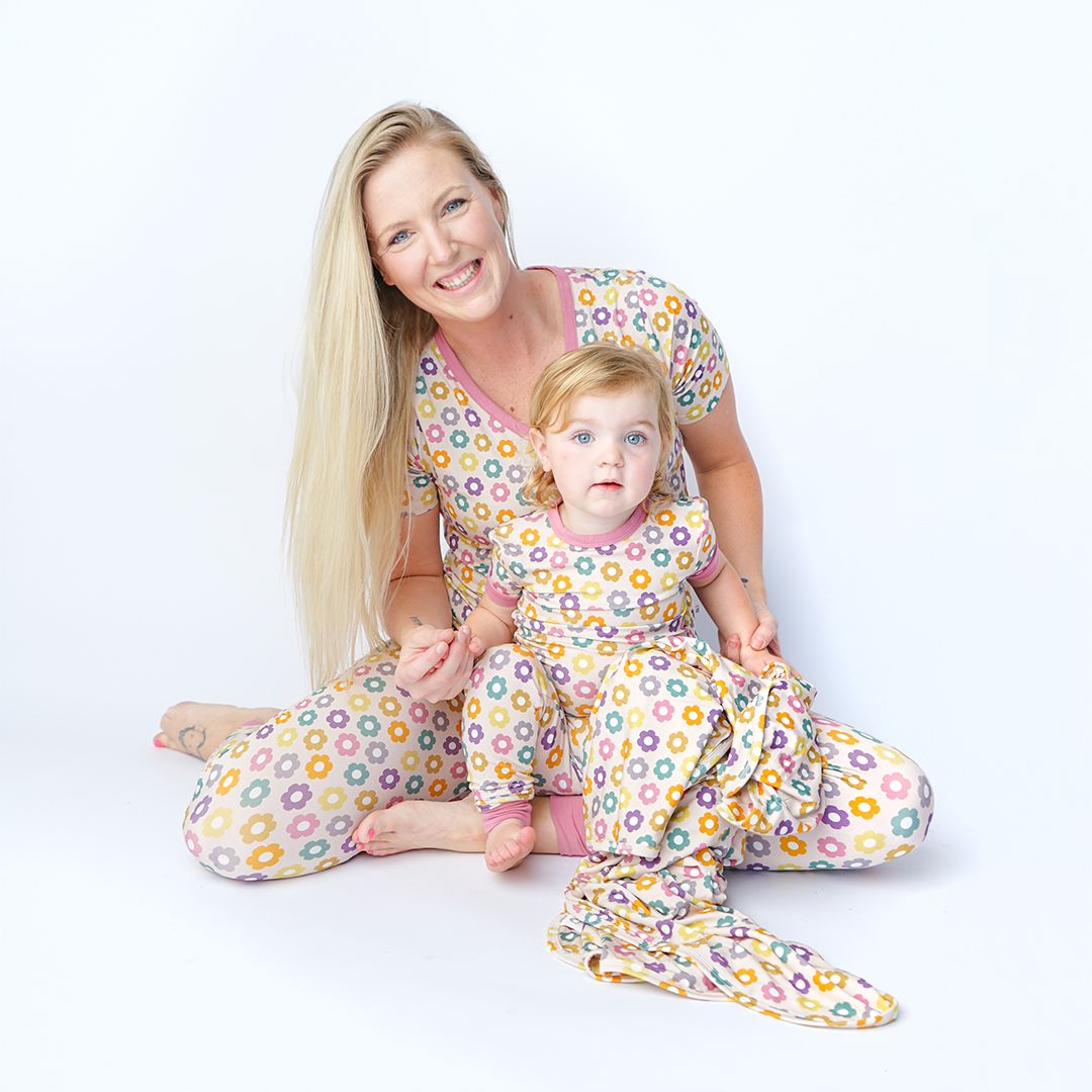 mom sits on the floor with her baby in her lap. they are all in the "feeling groovy" attire. mom wears the women's top and joggers. the baby wears the convertible. the "feeling groovy" print is a flowered print in multiple colors. these flowers are retro and groovy. the background is light pink. 