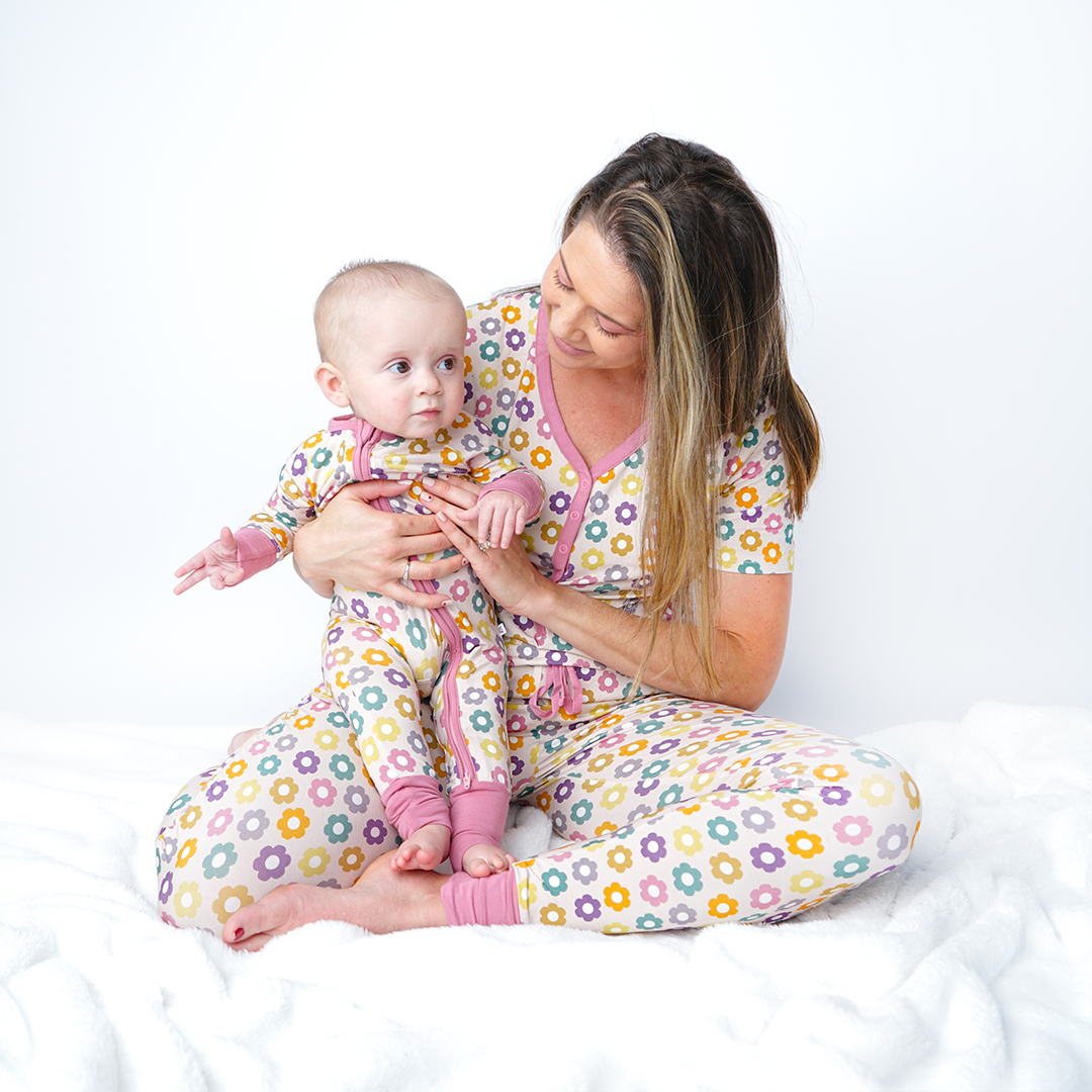 mom sits on the floor with her baby in her lap. they are all in the "feeling groovy" attire. mom wears the women's top and joggers. the baby wears the convertible. the "feeling groovy" print is a flowered print in multiple colors. these flowers are retro and groovy. the background is light pink. 