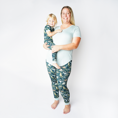 mom and daughter smile for the camera. the mom wears the "prehistoric friends" joggers and she hold her daughter on her hip . the daughter wears the 2-piece matching set. the "prehistoric friends" print is a mix of cute dinosaurs, eggs, and branches, all scattered on a dark green background.
