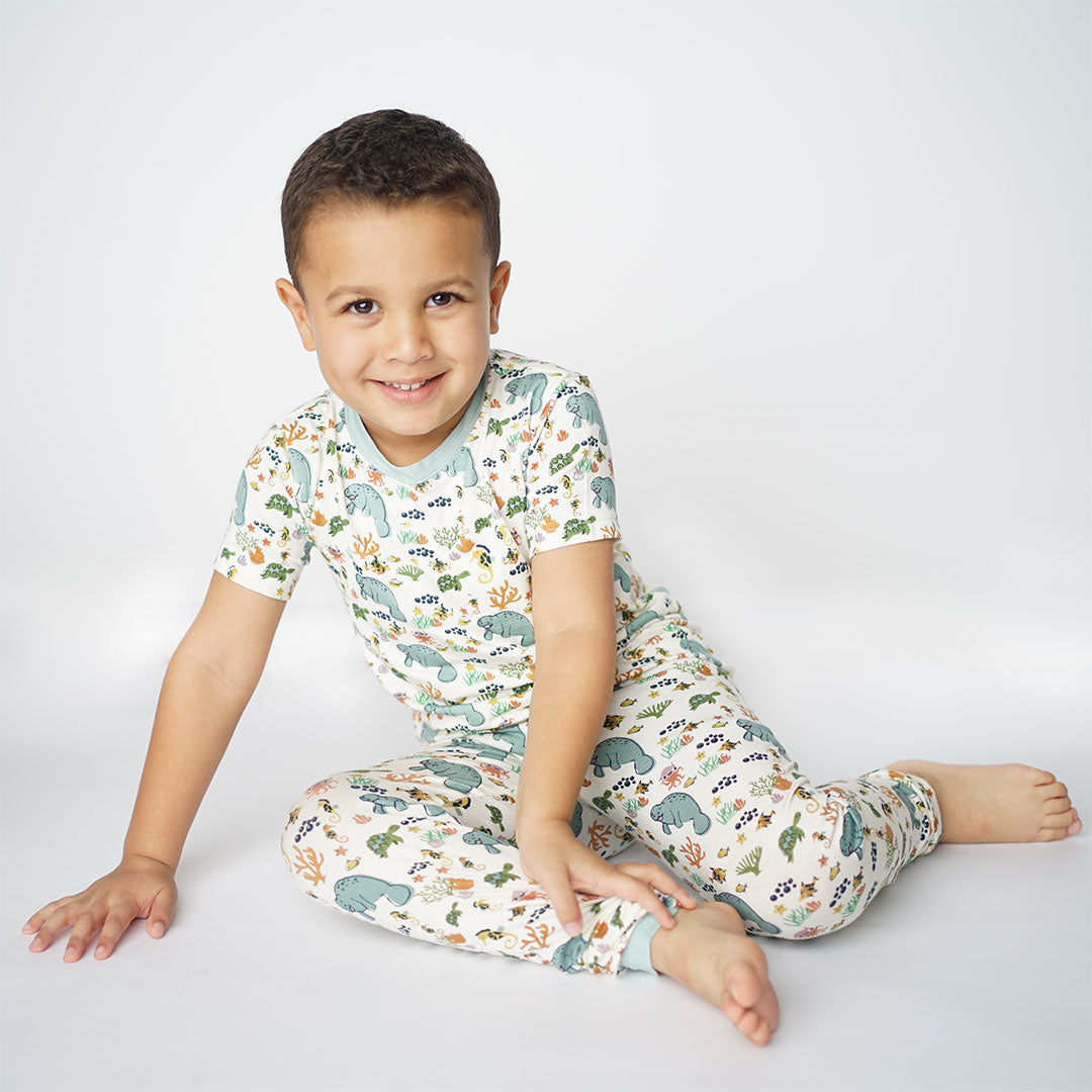 a boy sits on the floor posing. he is wearing the "manatee" 2-piece matching pajama set. the print "manatee" unites the florida sea life in a majestic print. there is a variation of florida manatees, yellow and pink sea horses, yellow and pink starfish, bubbles, turtles, and coral patterned across a white background