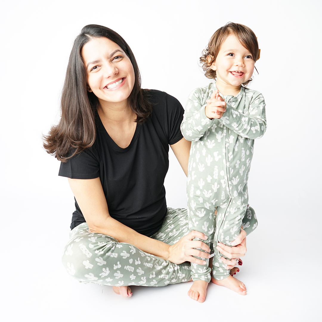 mom sits on the floor with her baby standing next to her. they both smile and wear "stay sharp" attire. the mom in the women's jogger and unisex black top. the baby wears the convertible. the "stay sharp" print is a variety of different white cacti on a greyish/green background. 