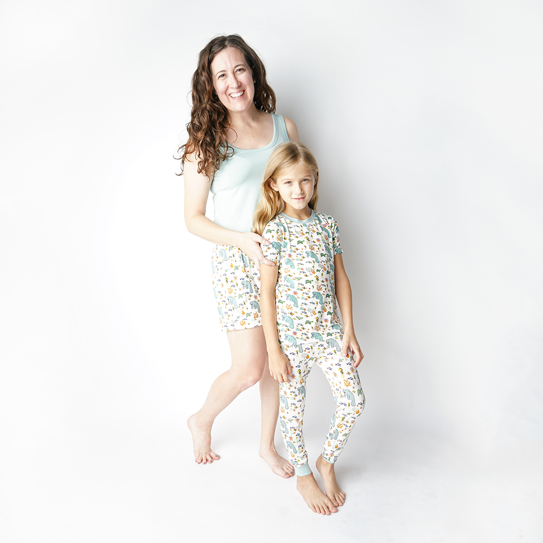 mom and daughter pose in their "manatee" apparel. the daughter stands with her 2-piece matching pajama set. the mom stands behind her in her "manatee" women's shorts and the "blue surf" tank top. the print "manatee" unites the florida sea life in a majestic print. there is a variation of florida manatees, yellow and pink sea horses, yellow and pink starfish, bubbles, turtles, and coral patterned across a white background