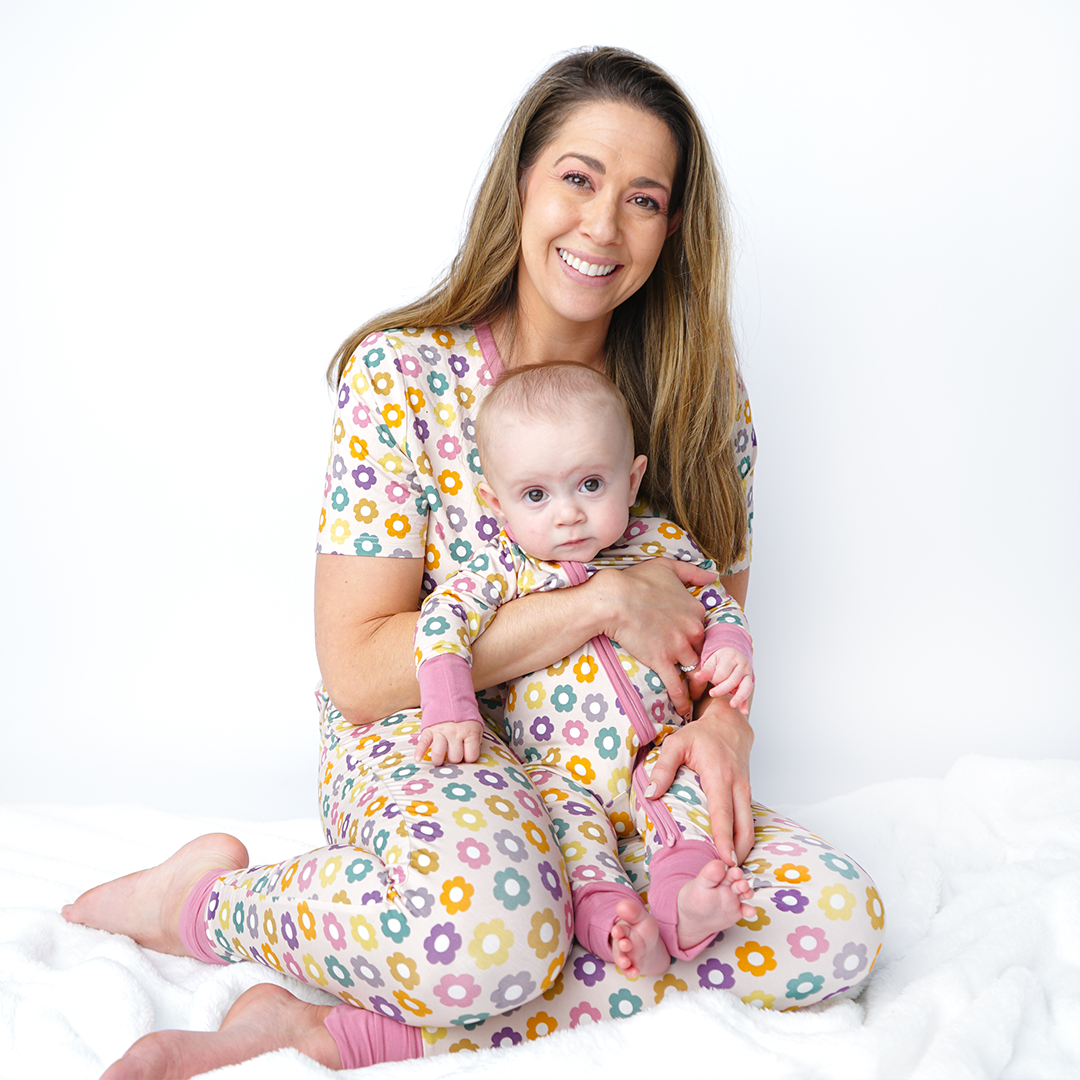 mom sits on the floor with her baby in her lap. they are all in the "feeling groovy" attire. mom wears the women's top and joggers. the baby wears the convertible. the "feeling groovy" print is a flowered print in multiple colors. these flowers are retro and groovy. the background is light pink. 