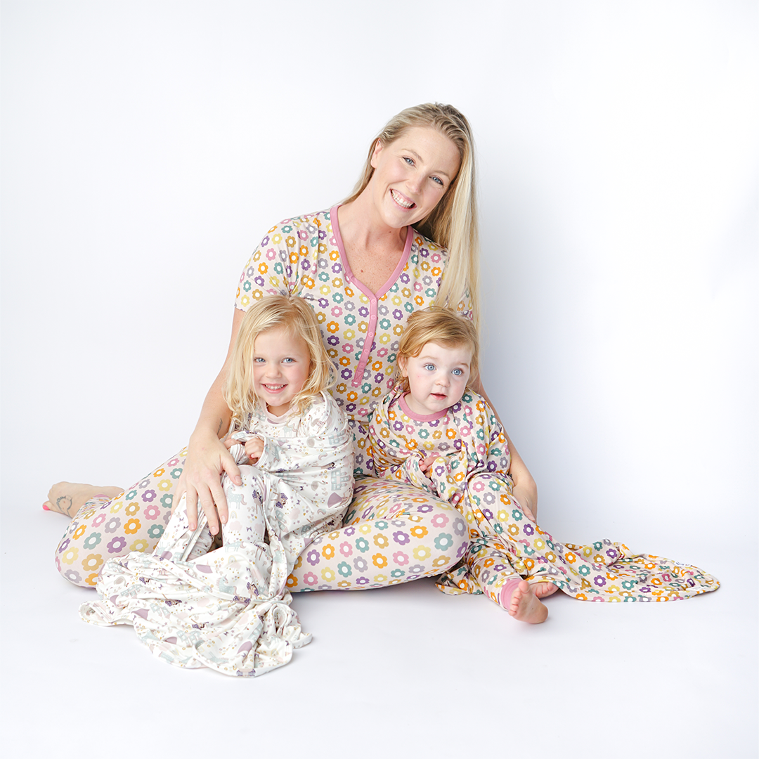 mom sits on the floor with her baby in her lap. they are all in the "feeling groovy" attire. mom wears the women's top and joggers. the baby wears the convertible. the "feeling groovy" print is a flowered print in multiple colors. these flowers are retro and groovy. the background is light pink. 