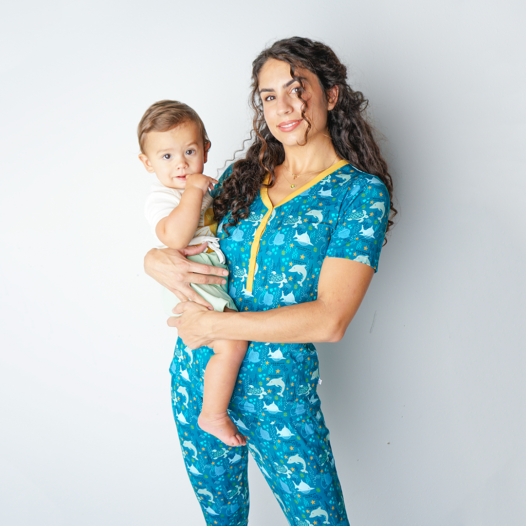 mom poses in her "ocean friends" women's top and joggers, while holding her baby on her hip. the "ocean friends" print is a combination of dolphins, stingrays, fish, starfish, coral, bubbles, and sharks, all spread out on a deep sea blue background.