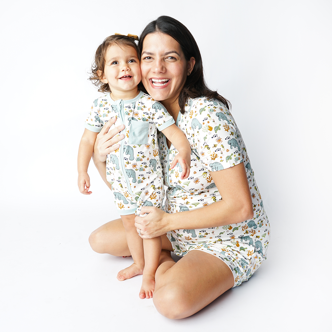 mom and daughter embrace one another while smiling at the camera. the mom wears the "manatee" women's top and women's shorts, while the daughter wears the "manatee" shortie. the print "manatee" unites the florida sea life in a majestic print. there is a variation of florida manatees, yellow and pink sea horses, yellow and pink starfish, bubbles, turtles, and coral patterned across a white background