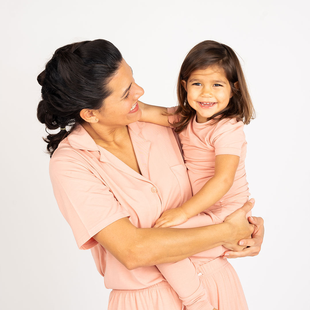 mom and daughter laugh at each other. the daughter is on her moms hip wearing the "dusty rose solid" matching 2-piece pajama set. the mom is wearing the "dusty rose solid" women's button down top and the women's shorts. 