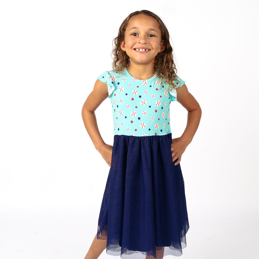 girl poses in her "baseball buddies" twirl dress. the "baseball buddies" print is a combination of baseballs, red stars, and blue stars scattered across a teal background. 