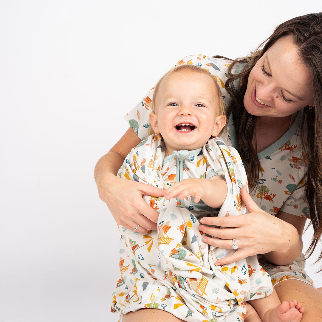 a mom smiles and sits on her knees with her baby cheerfully laughing. he is wrapped in the "beach days" blanket. he wears the "beach days" shortie romper. mom is wearing the "beach days" women's top and women's shorts. the "beach days" print is a scattered print of beach umbrellas, beach balls, sand buckets and shovels, coral, shells, beach chairs, seagulls, flipflops, and sunglass mixed in a orderly and creative way. 