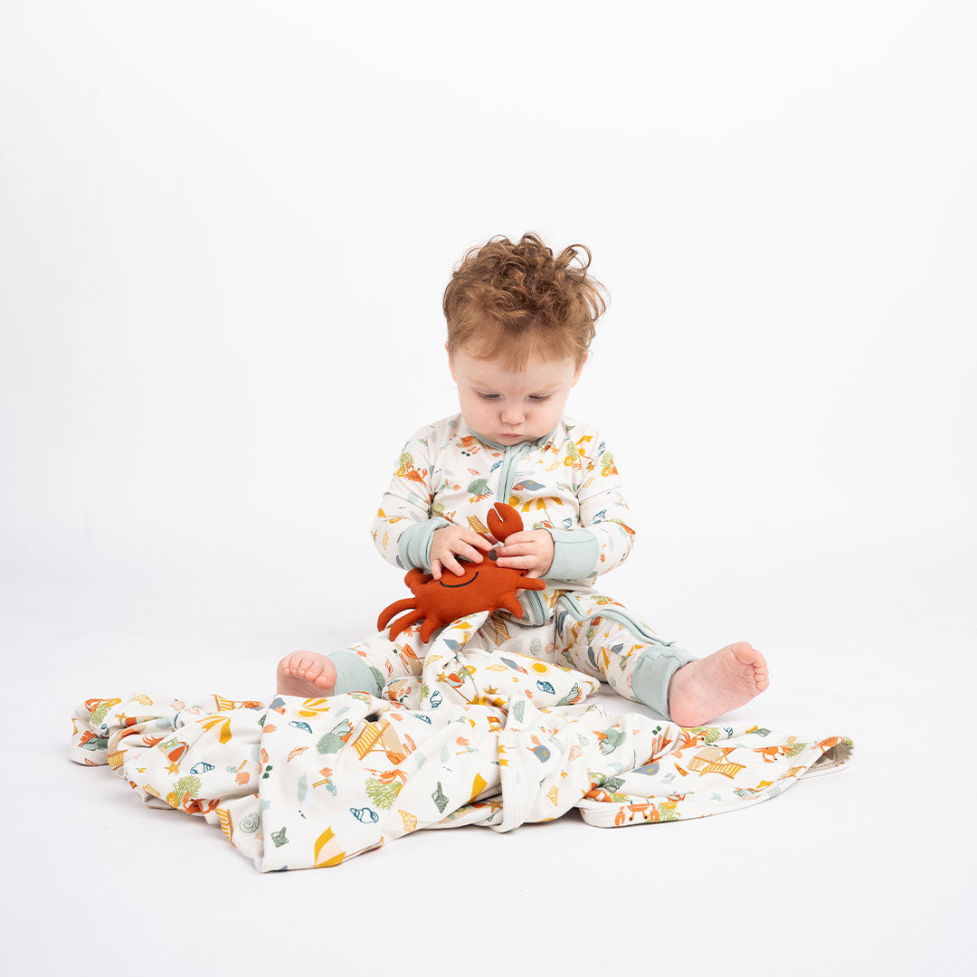 a baby sits on aa the "beach days" blanket, playing with his lovey. he is wearing the "beach days" convertible. the "beach days" print is a scattered print of beach umbrellas, beach balls, sand buckets and shovels, coral, shells, beach chairs, seagulls, flipflops, and sunglass mixed in a orderly and creative way. 