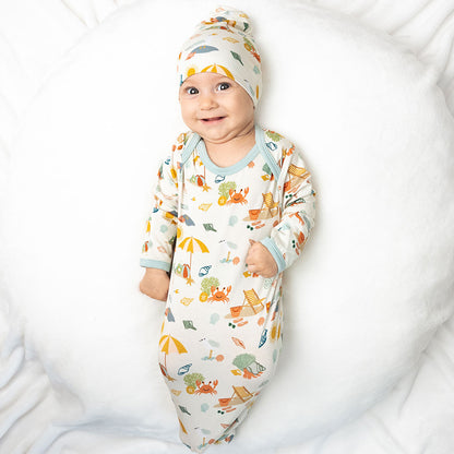 a baby smiles cheerfully in his "beach days" gown and hat set. he lays on his back on a white pillow. the "beach days" print is a scattered print of beach umbrellas, beach balls, sand buckets and shovels, coral, shells, beach chairs, seagulls, flipflops, and sunglass mixed in a orderly and creative way. 