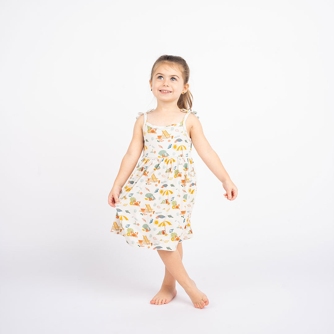 a girl stands with her legs crossed, arm is out to the side and the other one holds the side of her dress to show it off. she smiles, looking at the celling. she is wearing the "beach days" bamboo sundress. the "beach days" print is a scattered print of beach umbrellas, beach balls, sand buckets and shovels, coral, shells, beach chairs, seagulls, flipflops, and sunglass mixed in a orderly and creative way. 