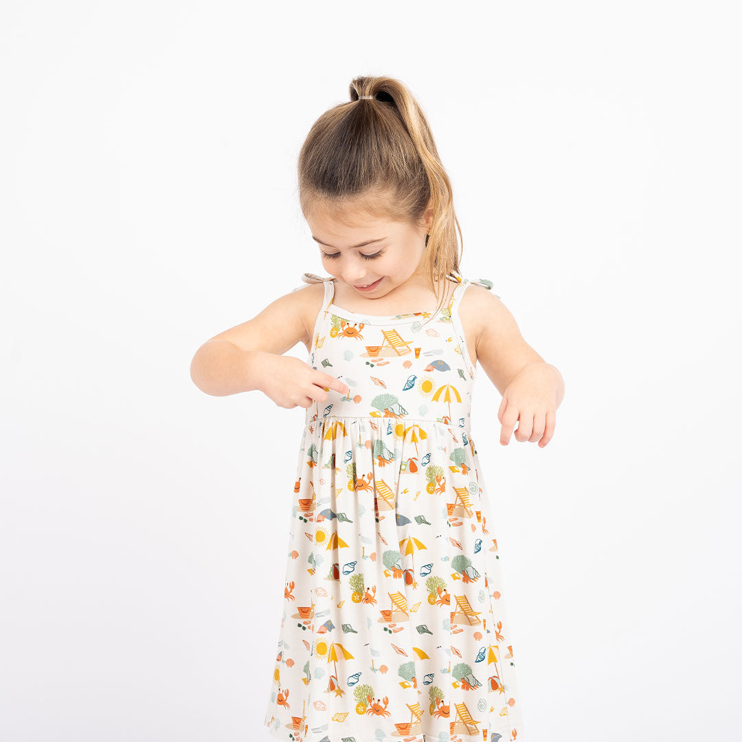 a young girl stands smiling and pointing at her "beach days" bamboo sundress. the "beach days" print is a scattered print of beach umbrellas, beach balls, sand buckets and shovels, coral, shells, beach chairs, seagulls, flipflops, and sunglass mixed in a orderly and creative way. 