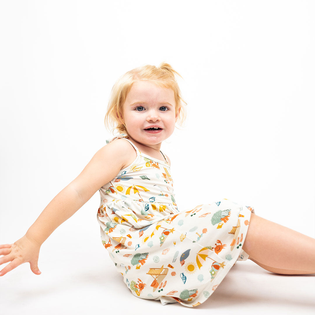 a little toddler girl sits on the floor with her body facing to the side. she smiles at the camera. she is wearing the "beach day" bamboo sundress. the "beach days" print is a scattered print of beach umbrellas, beach balls, sand buckets and shovels, coral, shells, beach chairs, seagulls, flipflops, and sunglass mixed in a orderly and creative way. 