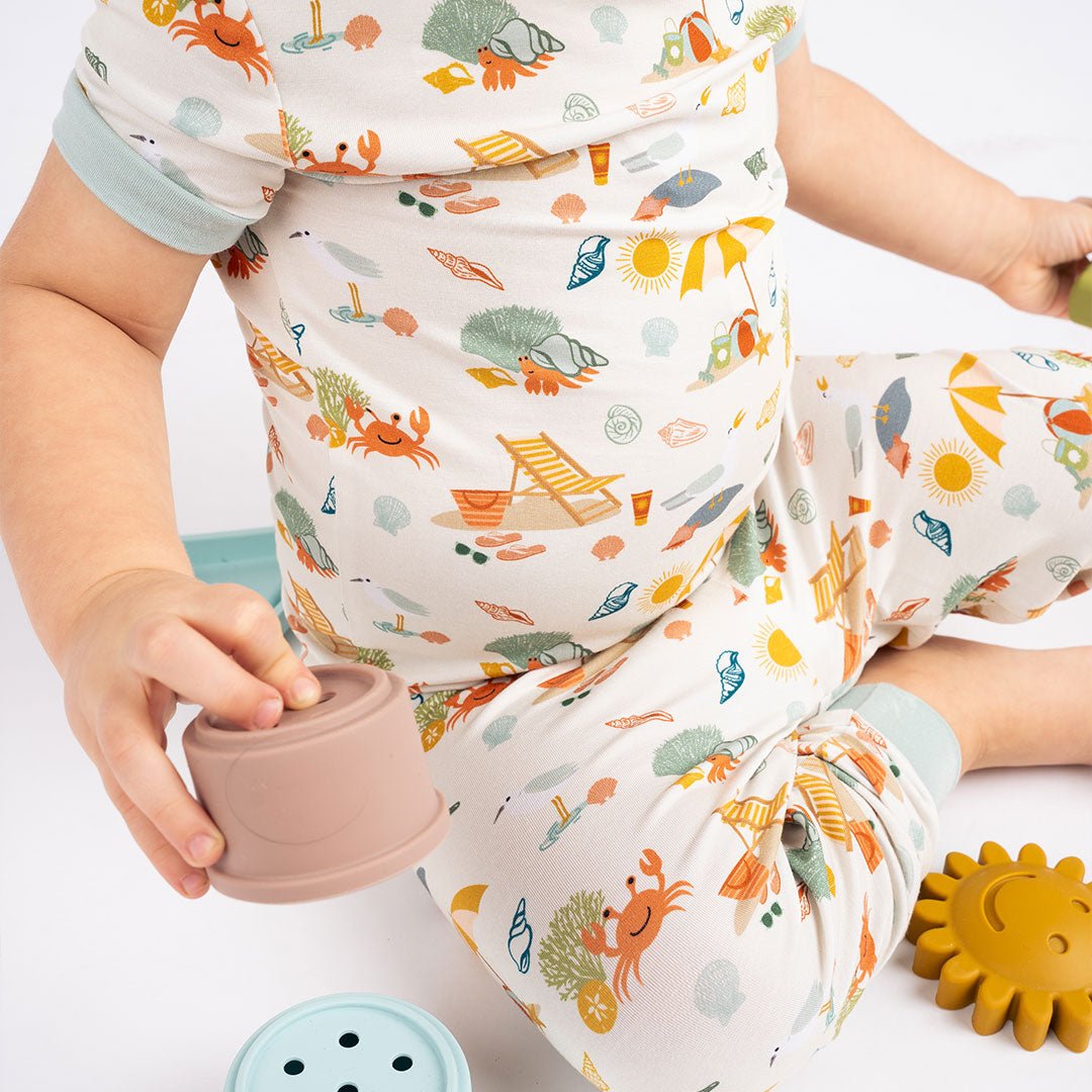 a baby sitting on the floor in his "beach days" 2-piece matching pajama set. he is playing with some silicone beach toys. the "beach days" print is a scattered print of beach umbrellas, beach balls, sand buckets and shovels, coral, shells, beach chairs, seagulls, flipflops, and sunglass mixed in a orderly and creative way. 