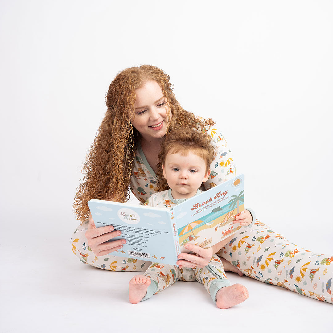 a mom reads the book "beach day" to her baby. they are wearing "beach days" prints. the mom wears the women's top and joggers. the baby is in the convertible. the "beach days" print is a scattered print of beach umbrellas, beach balls, sand buckets and shovels, coral, shells, beach chairs, seagulls, flipflops, and sunglass mixed in a orderly and creative way. 