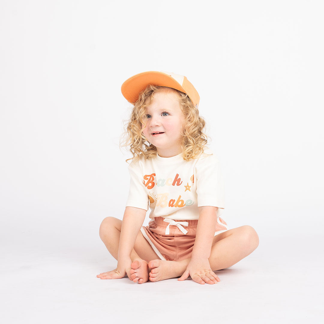 a girl sitting on the floor wearing the terry bottoms and the the "beach babe" cotton t-shirt. the word 'beach babe' is spelt out in a groovy font with a sun, starfish, and shell surrounding it. 