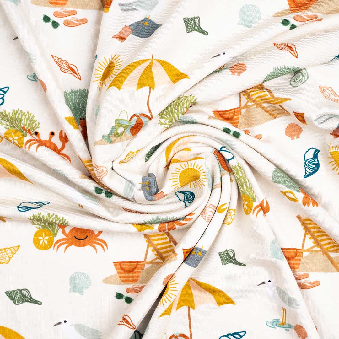 a photo of the blanket in a twirled up shape. the "beach days" print is a scattered print of beach umbrellas, beach balls, sand buckets and shovels, coral, shells, beach chairs, seagulls, flipflops, and sunglass mixed in a orderly and creative way. 