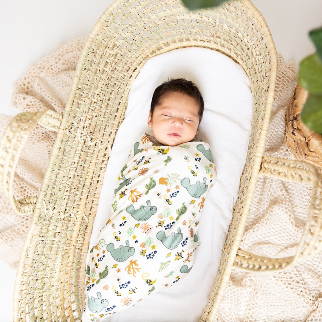 infant baby lays in a cradle on his back. he is wrapped up in the "manatee" bamboo blanket. the print "manatee" unites the florida sea life in a majestic print. there is a variation of florida manatees, yellow and pink sea horses, yellow and pink starfish, bubbles, turtles, and coral patterned across a white background